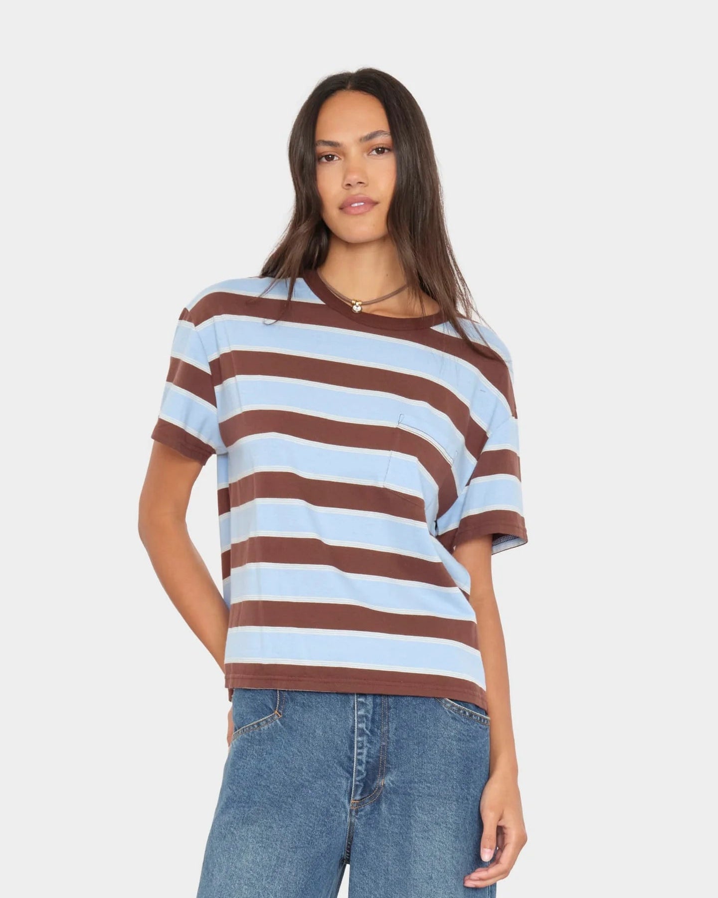 A woman modeling a short-sleeve crewneck t-shirt with horizontal brown and light blue stripes, paired with blue jeans, against a solid light gray background.