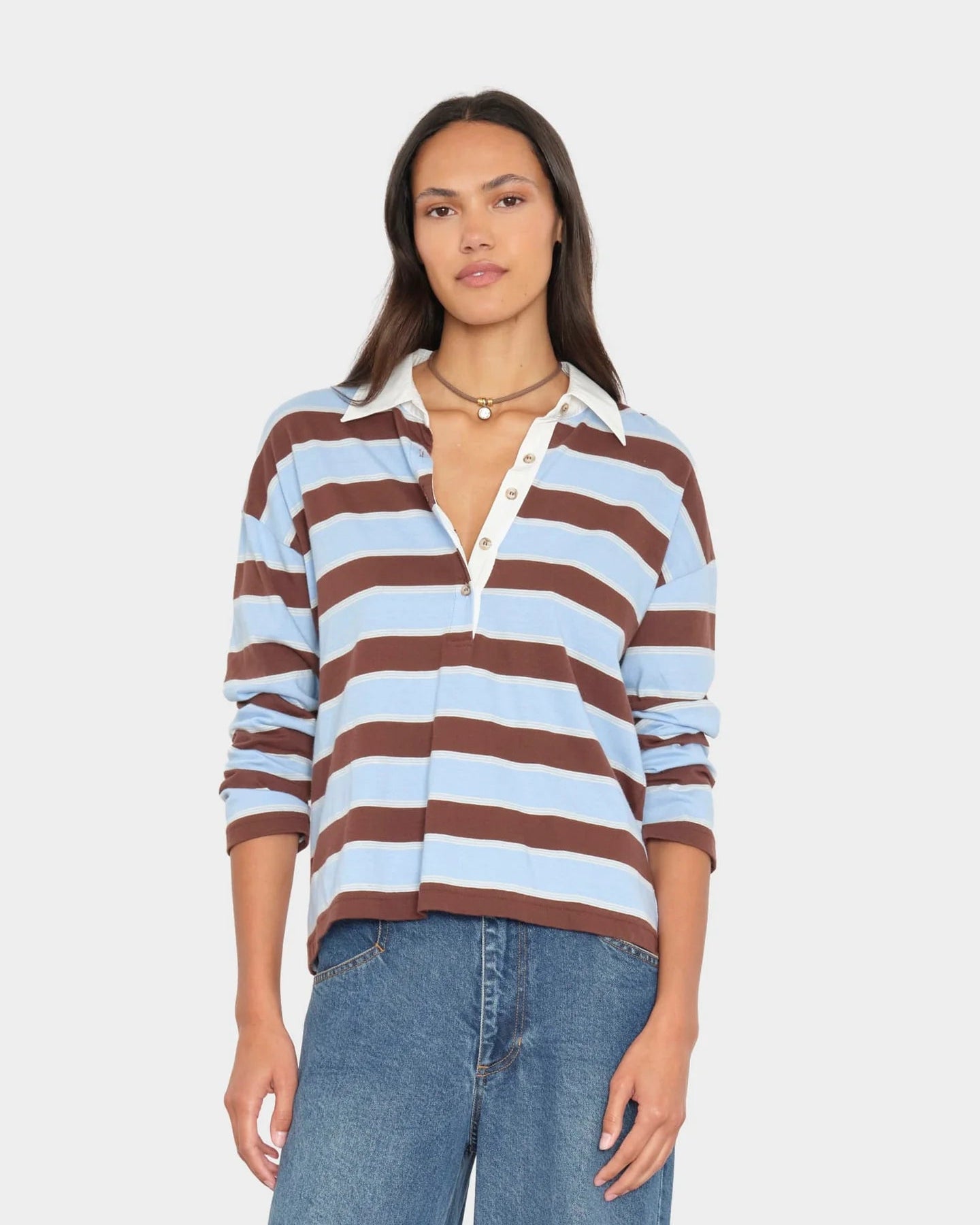 A woman modeling a long-sleeve knit polo-style top with horizontal brown and light blue stripes, a white collar, and a white button placket, paired with blue jeans against a light gray studio background.