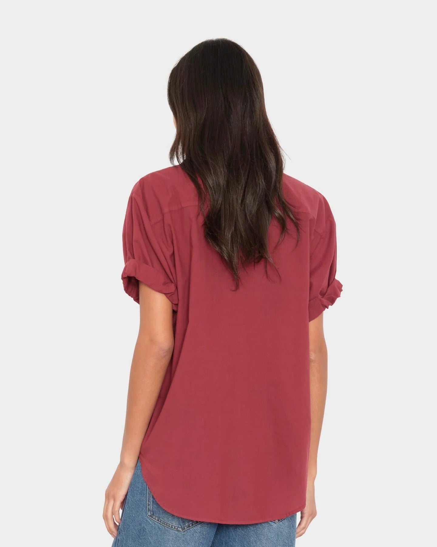 A woman viewed from behind wearing a muted red short-sleeve top with rolled cuffs and blue jeans, against a plain light gray studio background.
