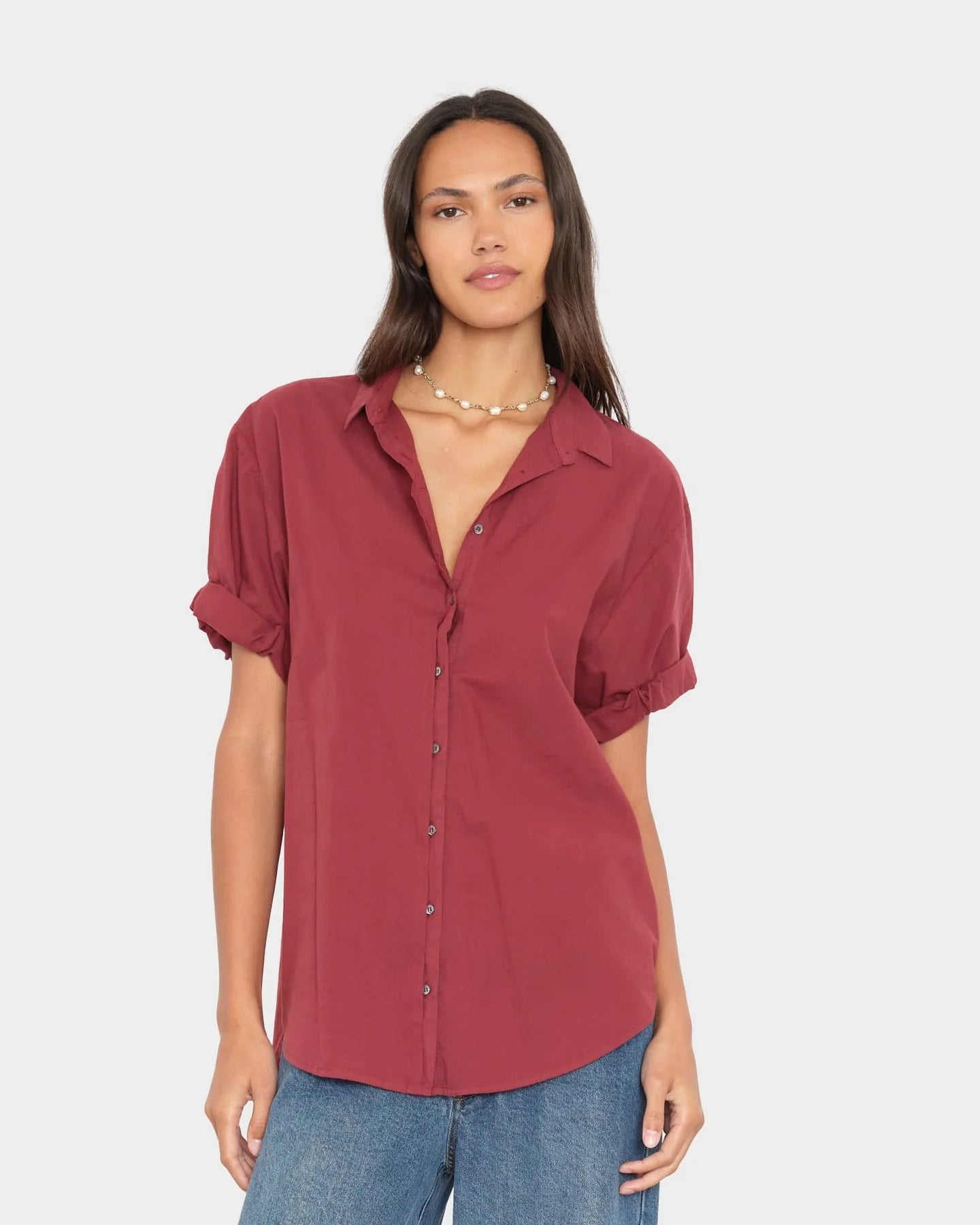 A woman modeling a short-sleeve button-up shirt in a dusty rose or muted burgundy color, paired with blue jeans, against a solid light gray background.
