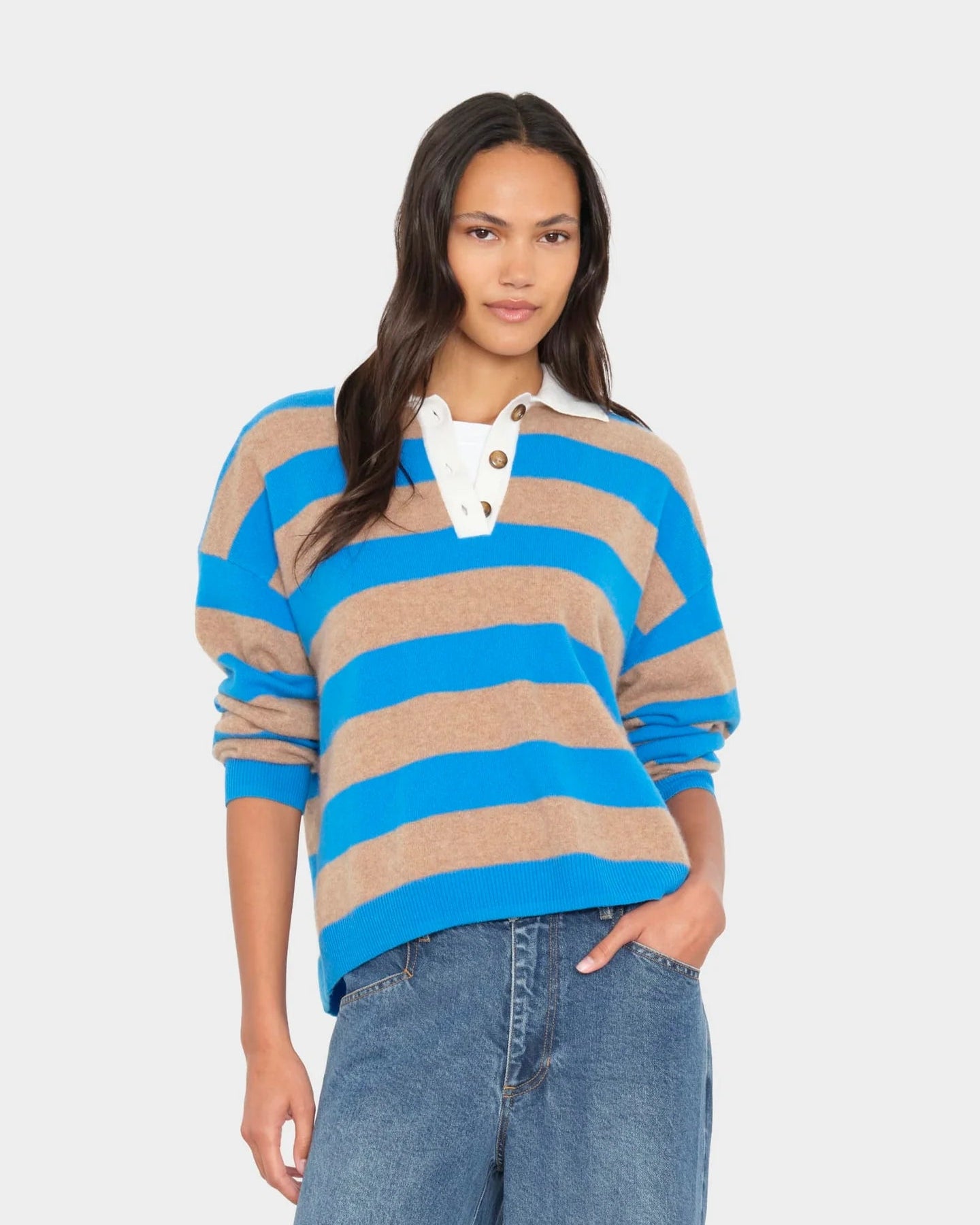 A woman modeling a blue and beige horizontally striped long-sleeve rugby sweater with a white collar and button placket, paired with blue jeans, against a light gray studio background.